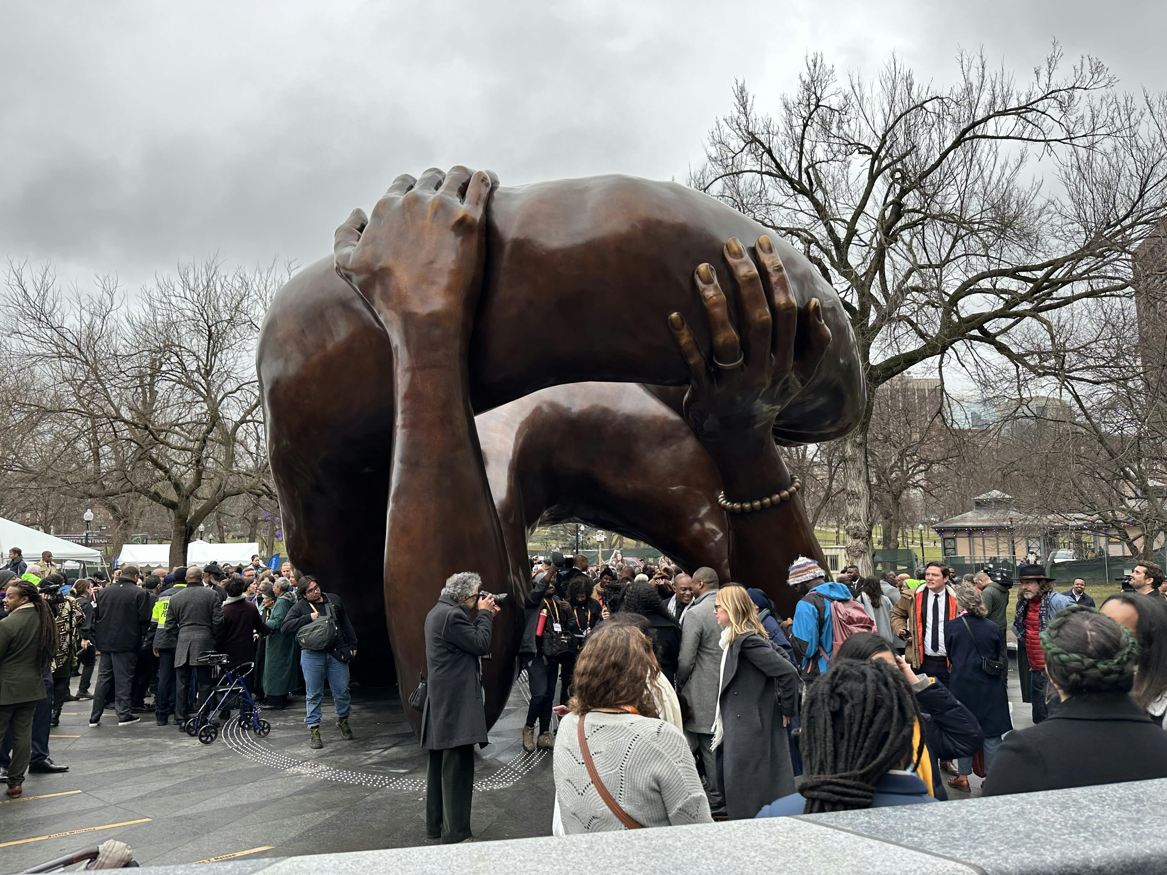 Large bronze sculpture of two hands holding up an indiscernible object, surrounded by a crowd of visitirs.