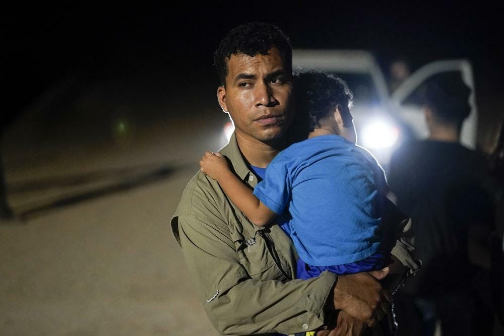 A Peruvian man wearing an olive green shirt holds his small child wearing a blue shirt. It is night time, and a cars headlights can be seen behind them.