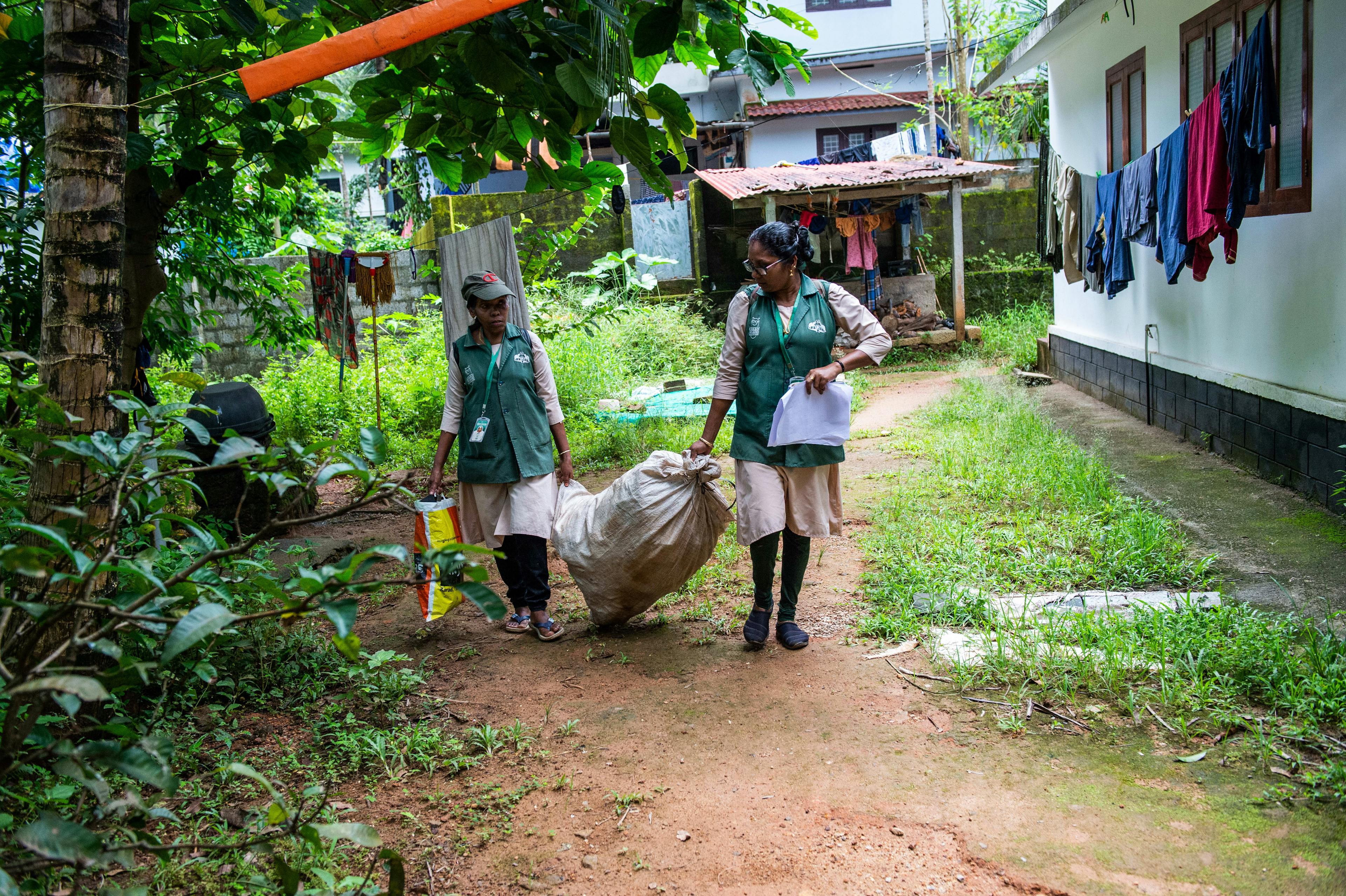 Two women wearing green vests and name tags around their neck carrying a white, full, large trash bag between them, walking outside next to a home. 