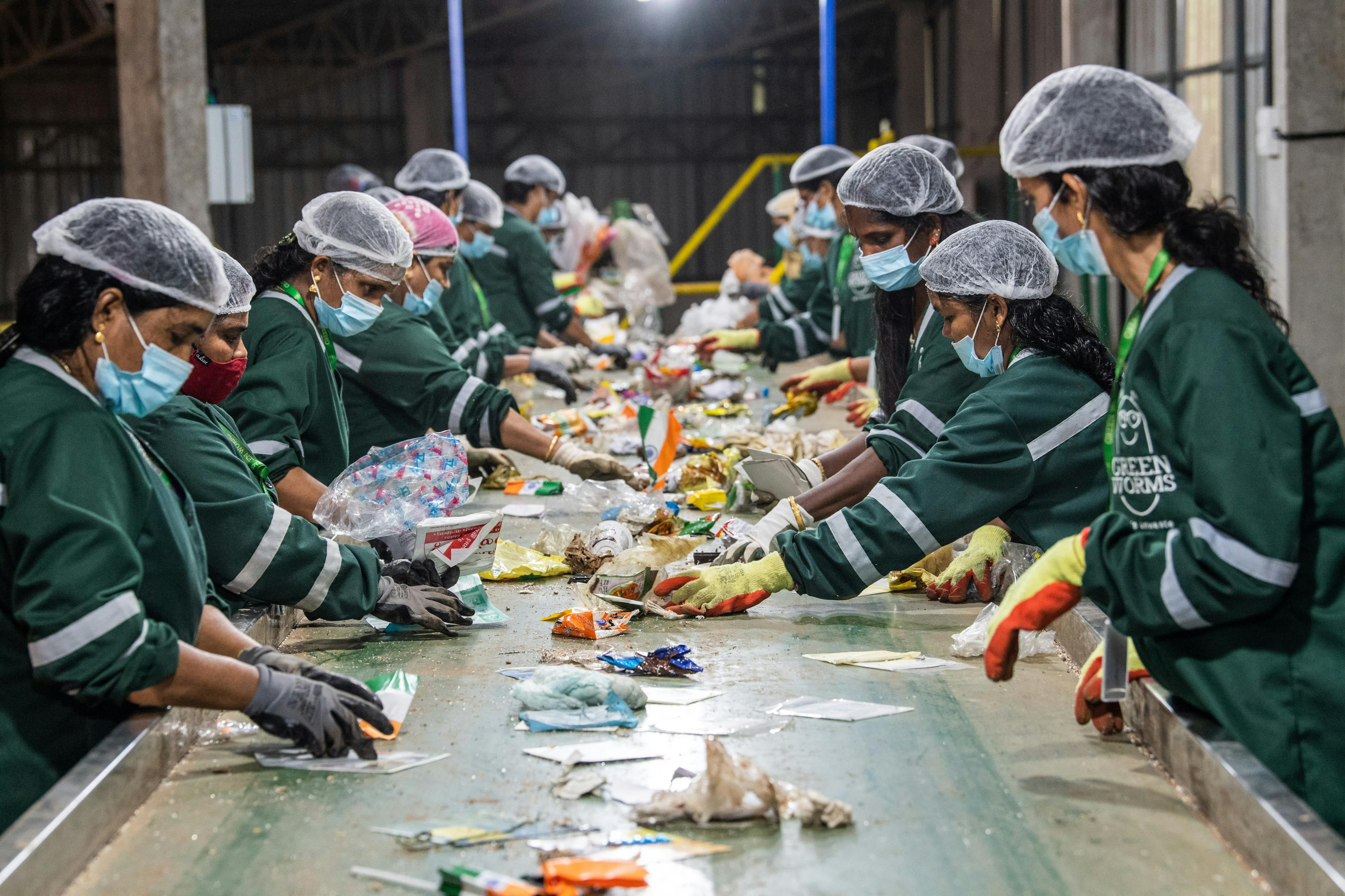 Looking over a sorting line with various pieces of waste on it. On each side, women wearing green uniforms and hair nets are sorting different pieces.