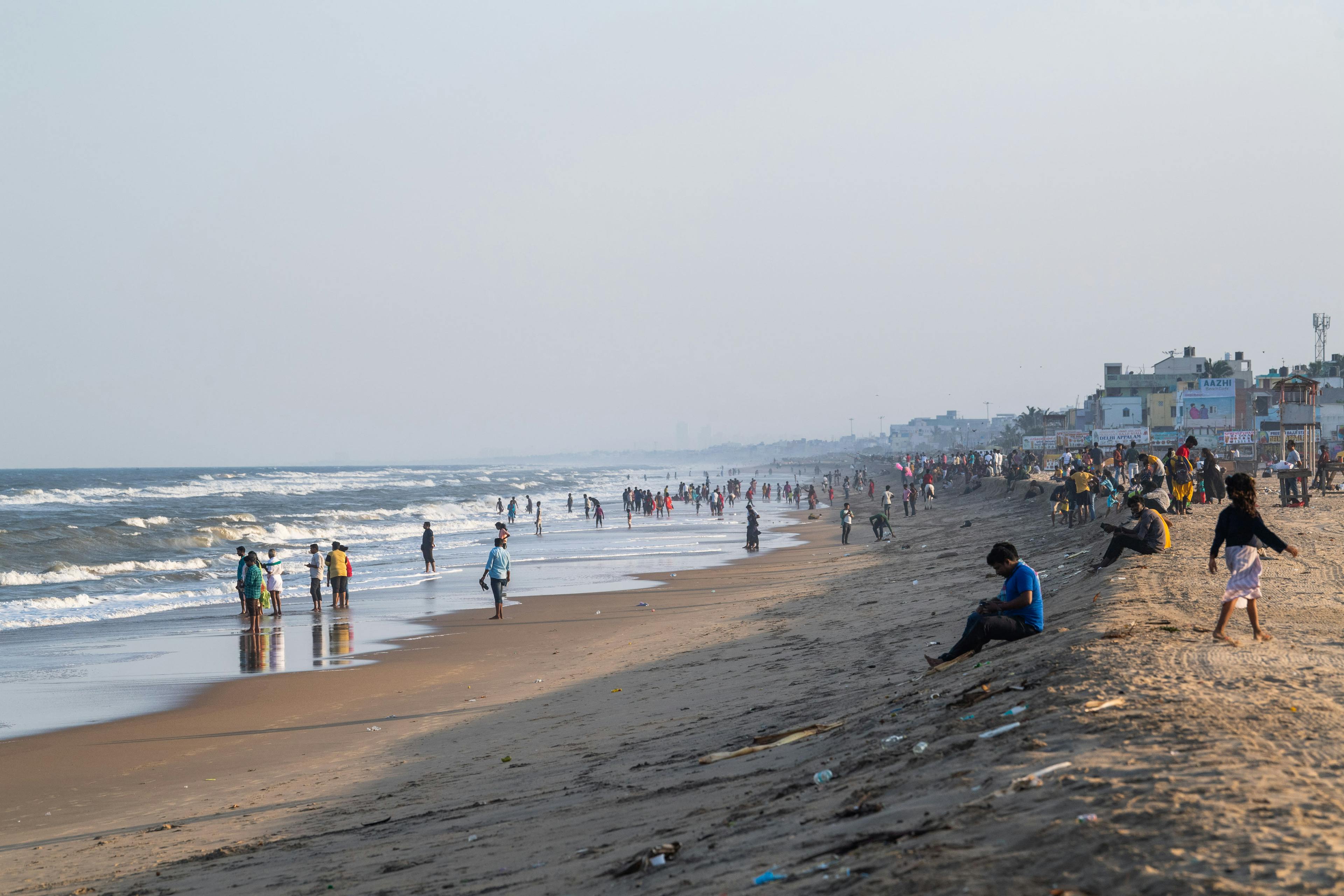 Daytime image of an overcast beach with people all around in India. A line of litter can be seen lining the beach.