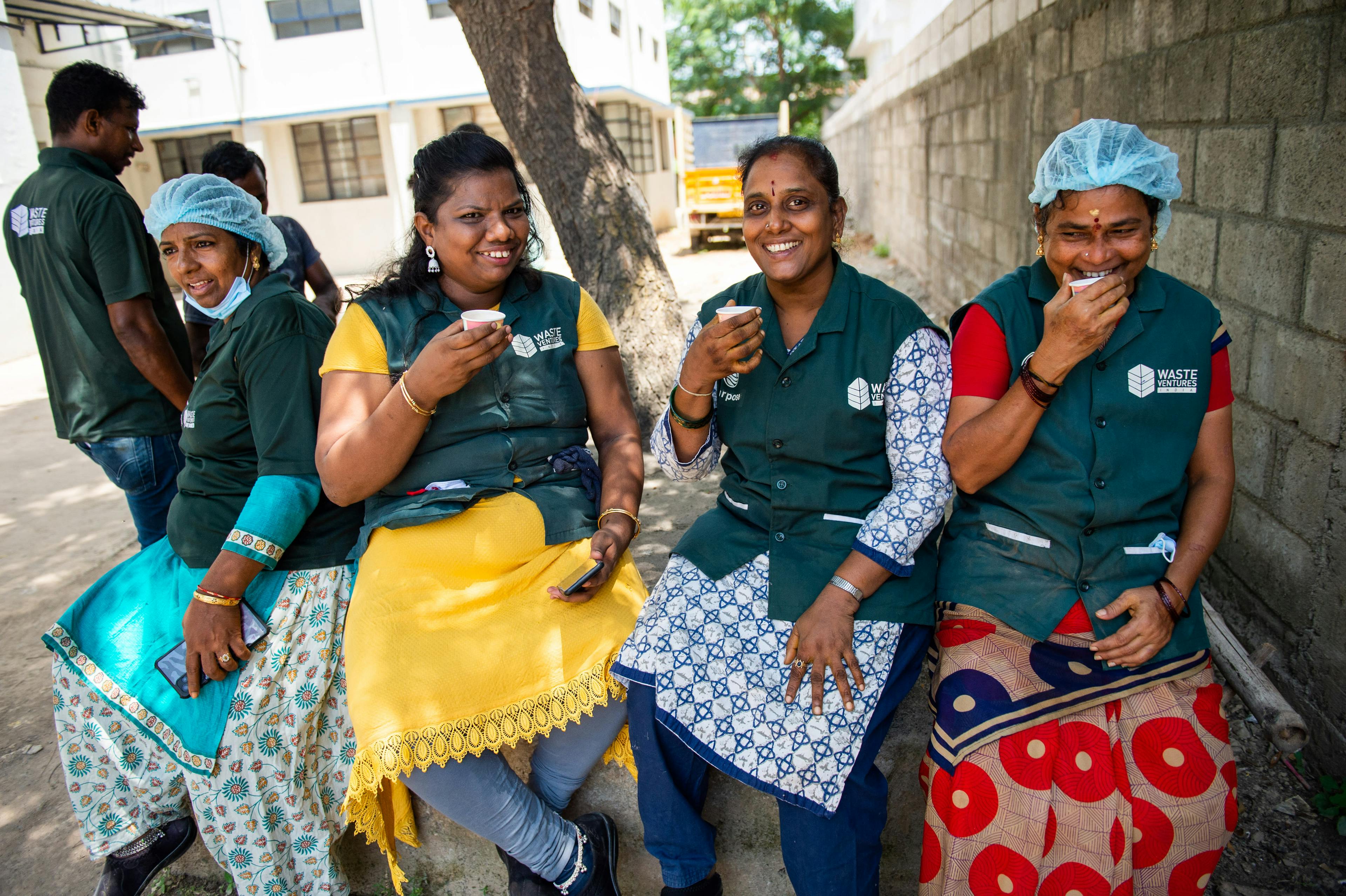 Four women sitting on break outside under shade, each wearing a green vest. They are smiling as they drink from a paper cup.