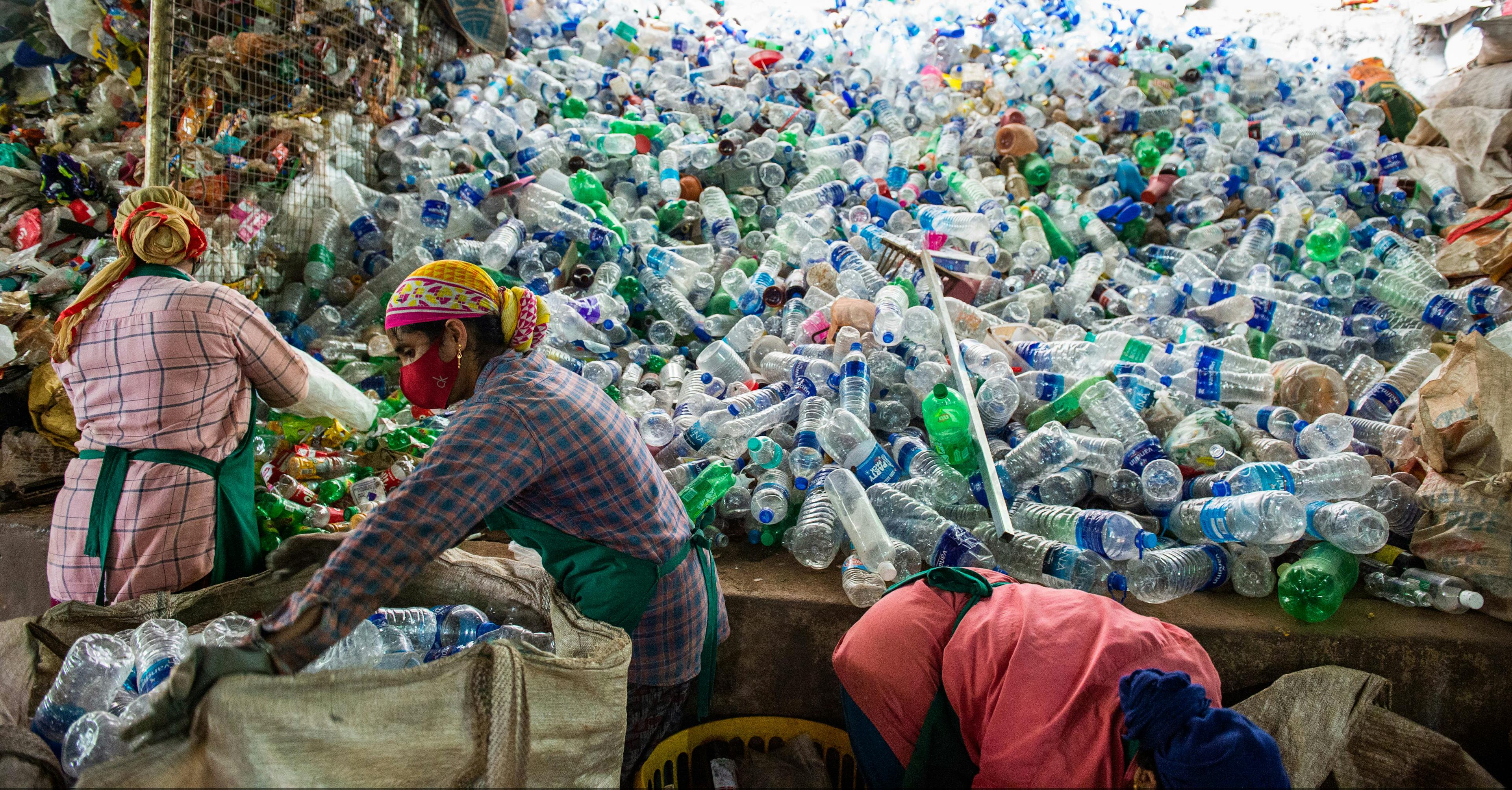 Two women are seen sitting on the lower right at the edge of a literal mountain of empty plastic bottles.