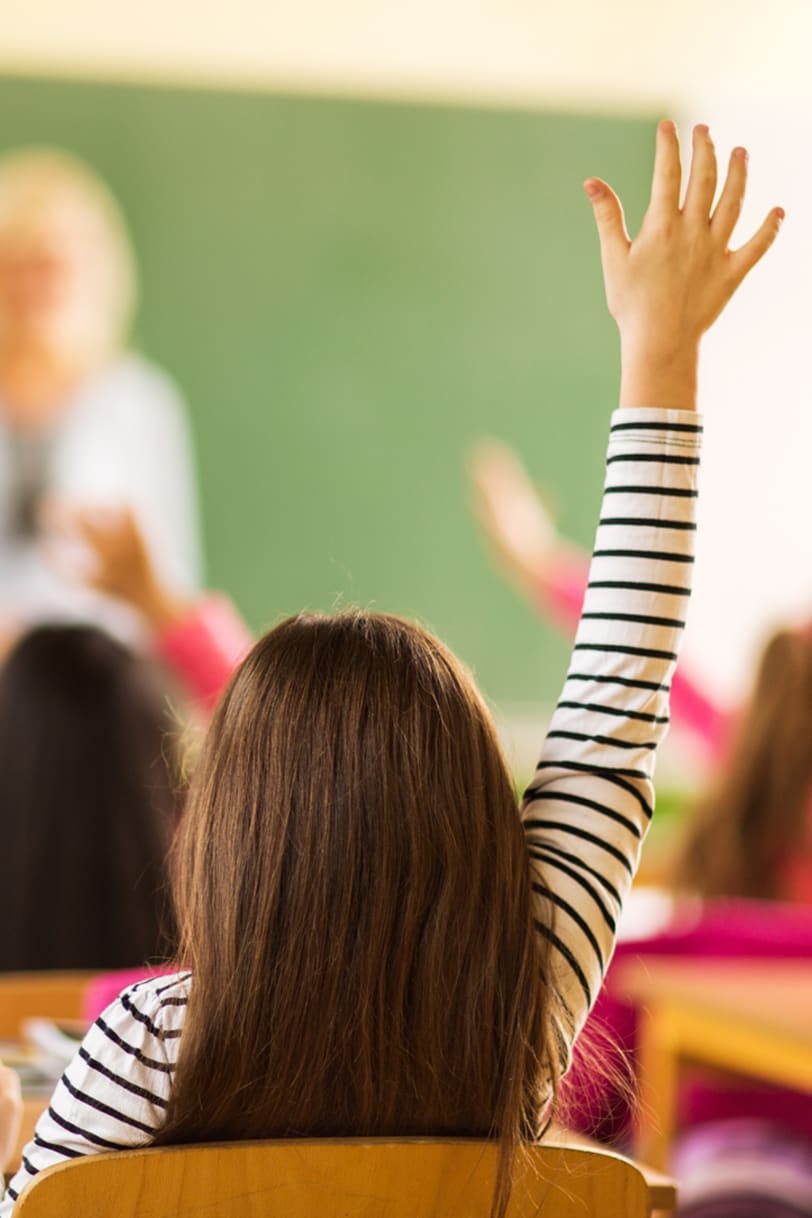 Child raising her hand in classroom, with the teacher in front of a blackboard in the background