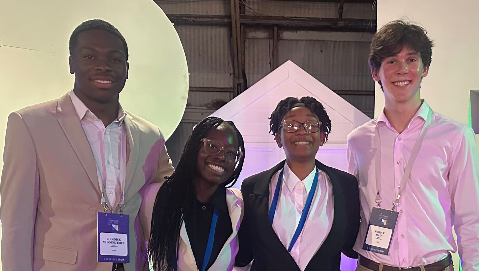 A dozen students standing together in 3 rows facing the camera taking a photo from the inside of a well lit building with a lot of glass panels